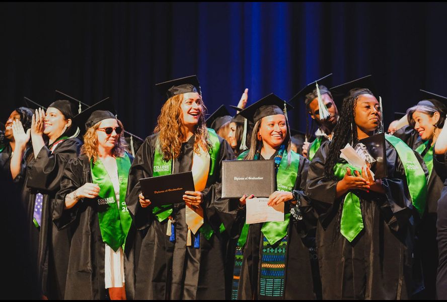 a group of students in black graduation robes with green stole smiling at their university commencement