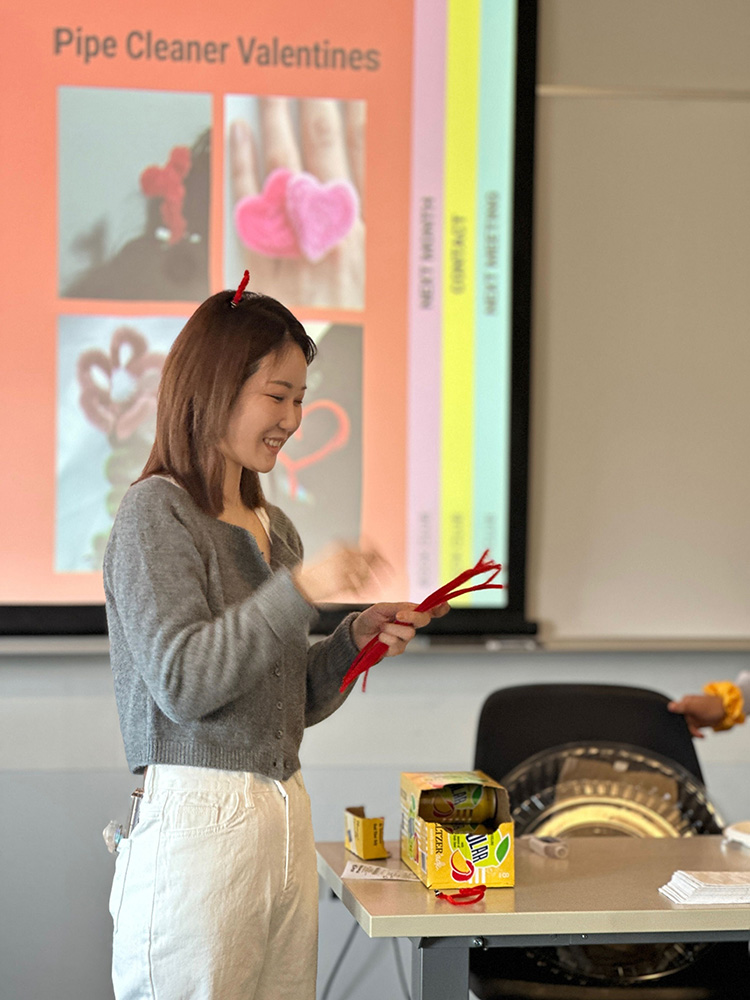 Jieun Lim (CFA'26), a Master of Arts in Art Education student at Boston University, wearing a gray sweater and white pants stands near a table, holding red pipe cleaners. On the table, there is a colorful box with more pipe cleaners and some other small items scattered around. Behind the person, a presentation slide is projected on the wall, titled "Pipe Cleaner Valentines," displaying images of heart-shaped pipe cleaner crafts. The setting appears to be a classroom or workshop environment.