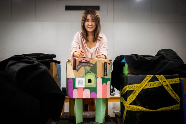 Jieun Lim (CFA'26), a Master of Arts in Art Education student at Boston University, is standing behind a colorful cardboard structure with four green legs, decorated with various paper cutouts and a QR code. Jieun appears to be working on or adjusting something on top of the structure. To the left and right of the structure are other cardboard creations partially covered with black fabric; the one on the right is wrapped with yellow caution tape. The setting appears to be an indoor room with a plain backdrop.