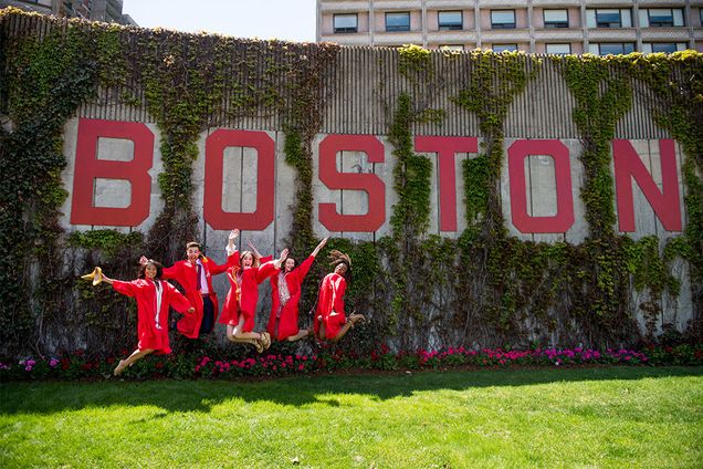 5 members of Boston University Class of 2019 wearing their red graduation robes jump in the air in front of the word Boston on Nickerson Field
