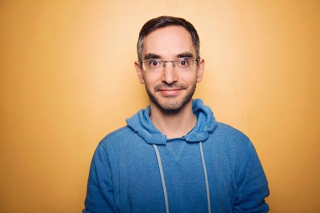 Portrait of Myq Kaplan. He wears a blue sweatshirt in front of an orange background.