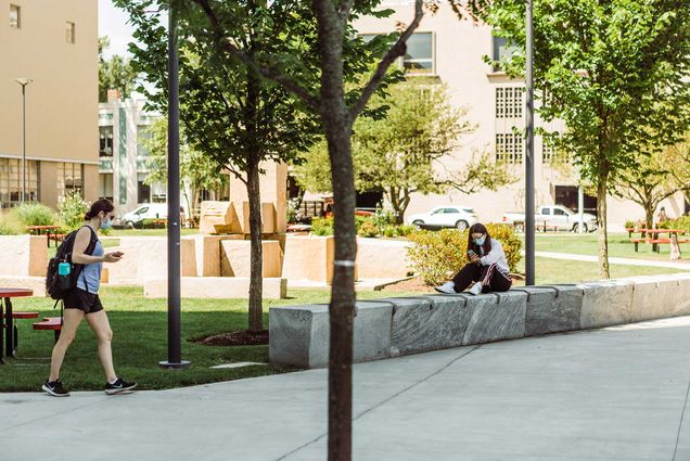 Photo of two students outside of Covid-19 testing lab at Kilachand Center with masks on. One sits on a bench and looks at their phone, the other walks by with a backpack.