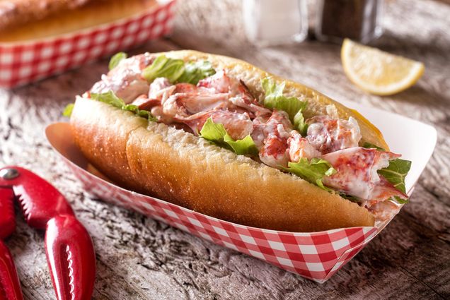 Photo of a lobster roll in a red and white checkerboard to-go container. To the left sits a lobster cracker in the form of two lobster claws.