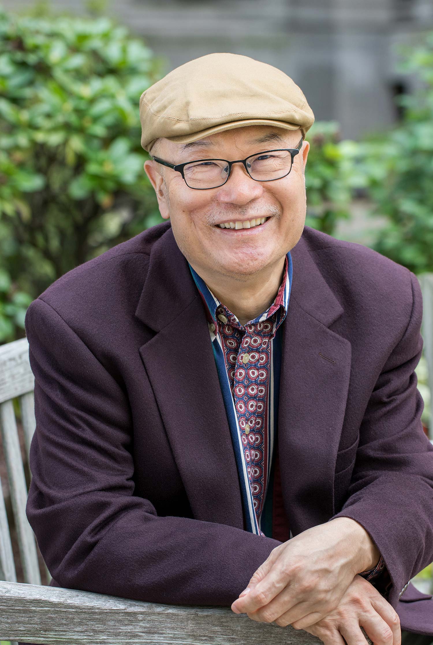 photo of Ha Jin in a tan scally cap and purpleish suit with a red, white and blue polka dot pattern tie. He sits on the edge of a park bench and crosses his hand over the arm rest.