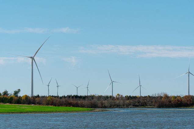 Photo of about a half dozen wind turbines. A body of water is seen in the foreground and some trees are seen in the background.