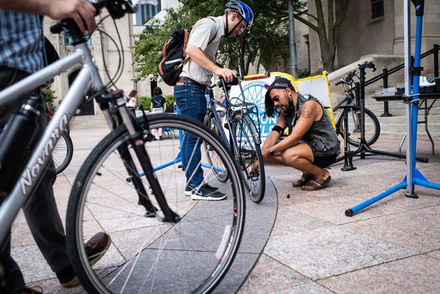 Photo of Teddy Heinrich, wearing a blue helmet and holding his bike, while Tristan Djaafar, crouching, in black, fixes something near his wheel. Another person holding their bike is seen in the left foreground. They appear to be on Marsh Plaza.