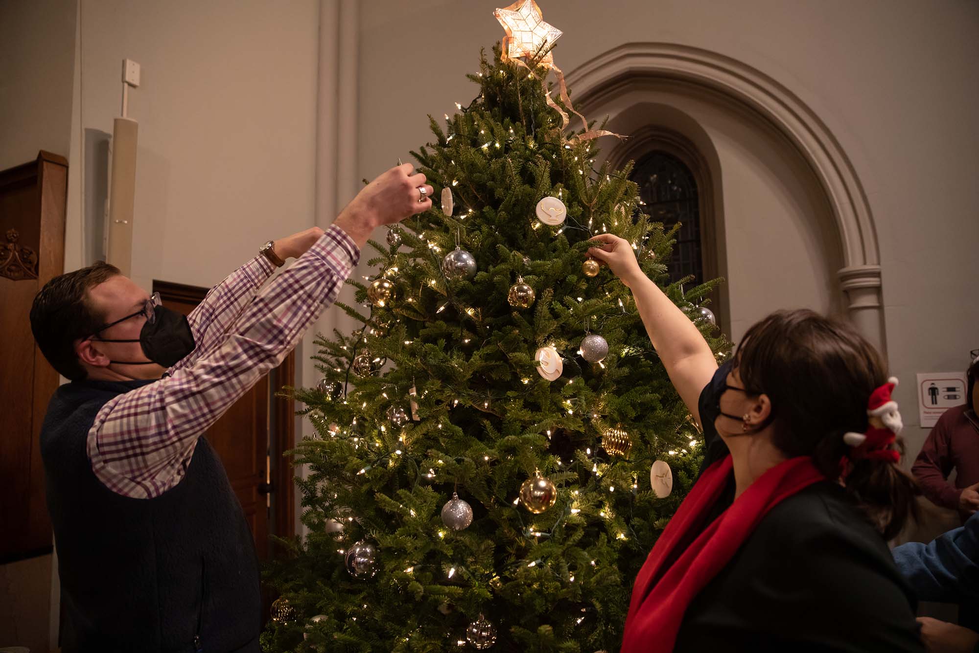 Photo of Clayton McCleskey (STH’24), left, and Sharon Solomon. (CAS’14) put the finishing touches on the Marsh Chapel Christmas tree. Both wear face masks. The tree is decorated with yellow lights and cream-colored ornaments, and has a five-pointed star on top.