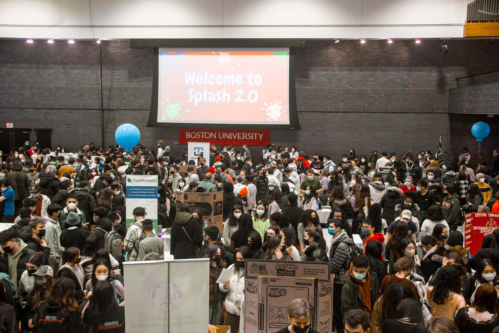Photo of Splash 2.0, the Club Fair at the beginning of the second semester each school year, is held at GSU on Saturday, January 22, 2022. The GSU is filled with students, masked, many wearing winter coats, who stand and talk with students tabling with trifold boards and signs. On the back wall, "Welcome to Splash 2.0" is projected.