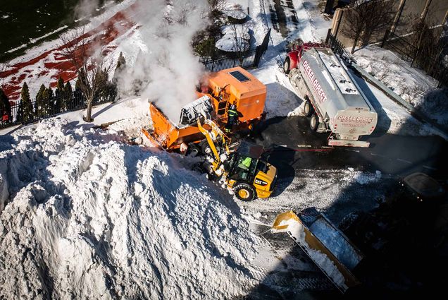 an aerial photo of construction equipment moving snow into a snow-melting machine along Agganis Way. A diesel truck sits nearby at the ready and a snow-covered soccer field is seen in the distance.