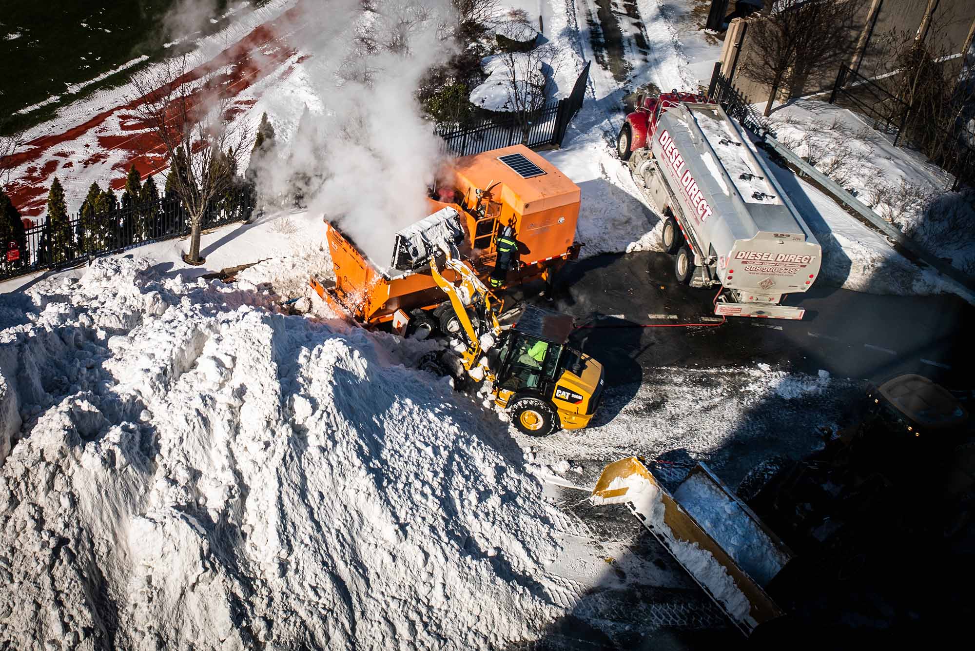 an aerial photo of construction equipment moving snow into a snow-melting machine along Agganis Way. A diesel truck sits nearby at the ready and a snow-covered soccer field is seen in the distance.