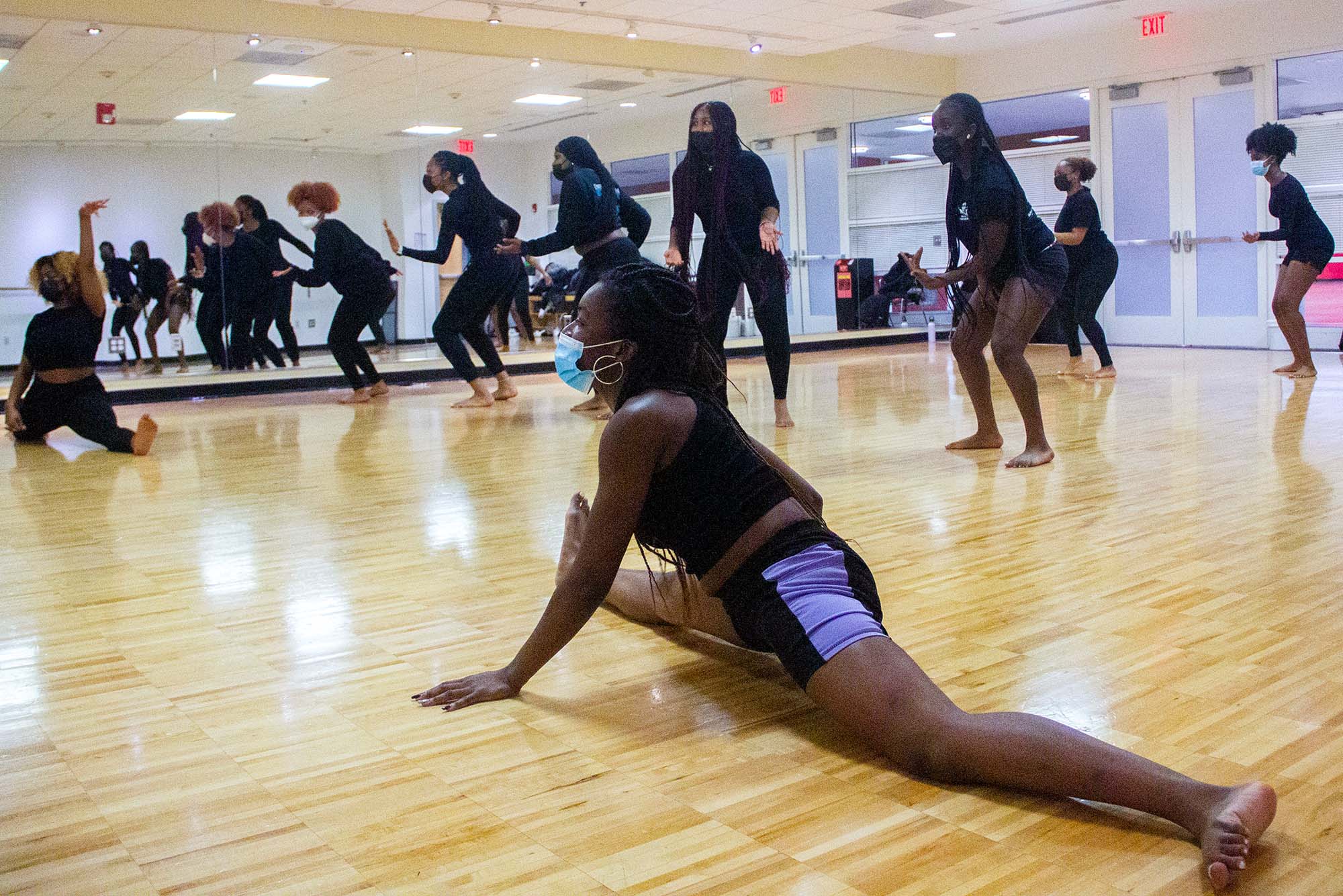 Photo of BU’s African Students Organization rehearsing at FitRec Feb 16 in preparation for Northeastern African Student Organization’s annual Africa Night. A young Black woman with braids and wearing a black athletic outfit does a split in the foreground as dancers dressed is black behind her do a sort of squat move. All the women wear masks. They are dancing in a studio with wooden floor and a wall-length mirror on the back wall.