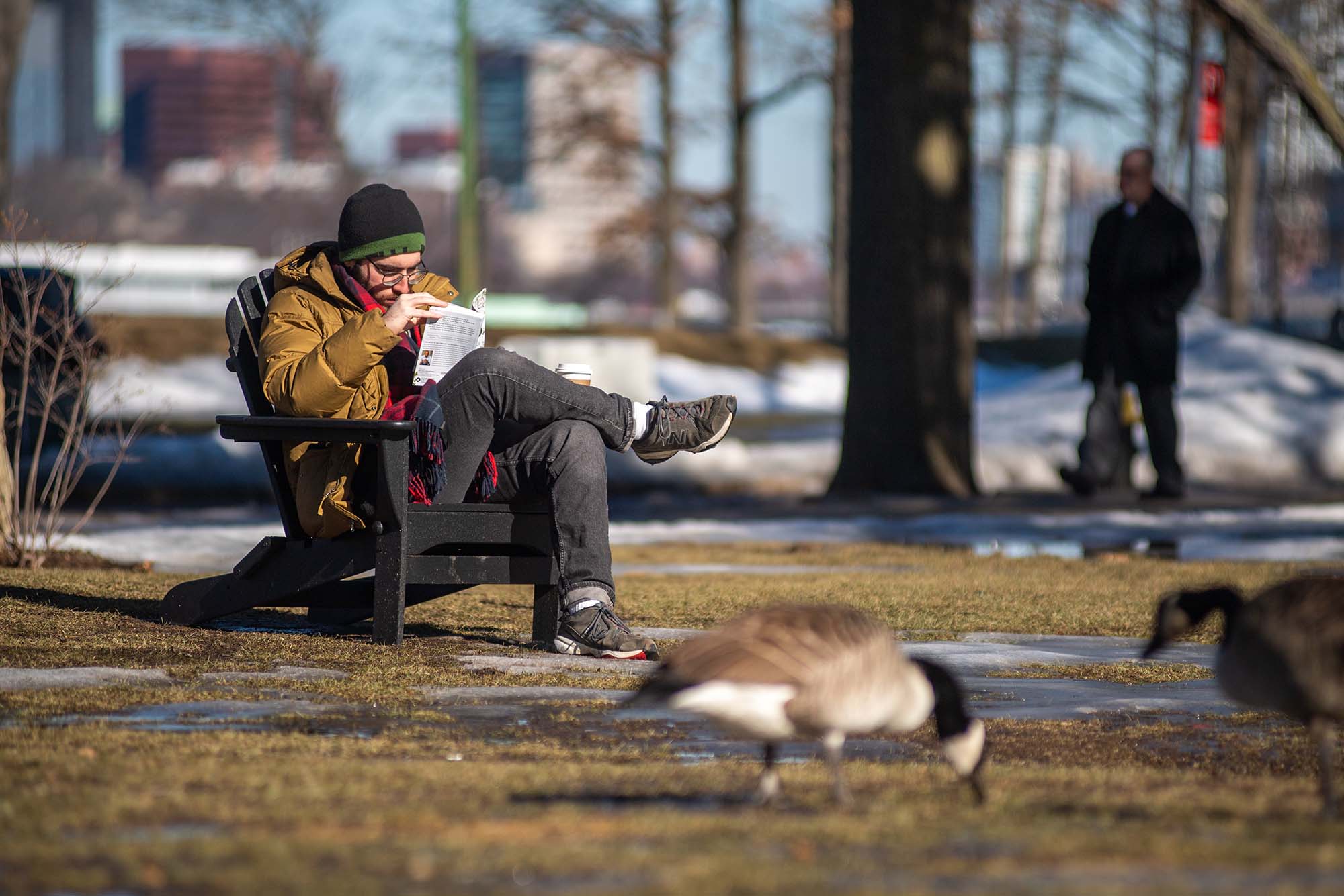 Photo of Tom Fischer (LAW’25,L1) digging into the book Just Mercy as he sits on a black Adirondack chair on BU Beach. He wears a gold puffy, black pants and a hat. A couple of geese are seen in the foreground.