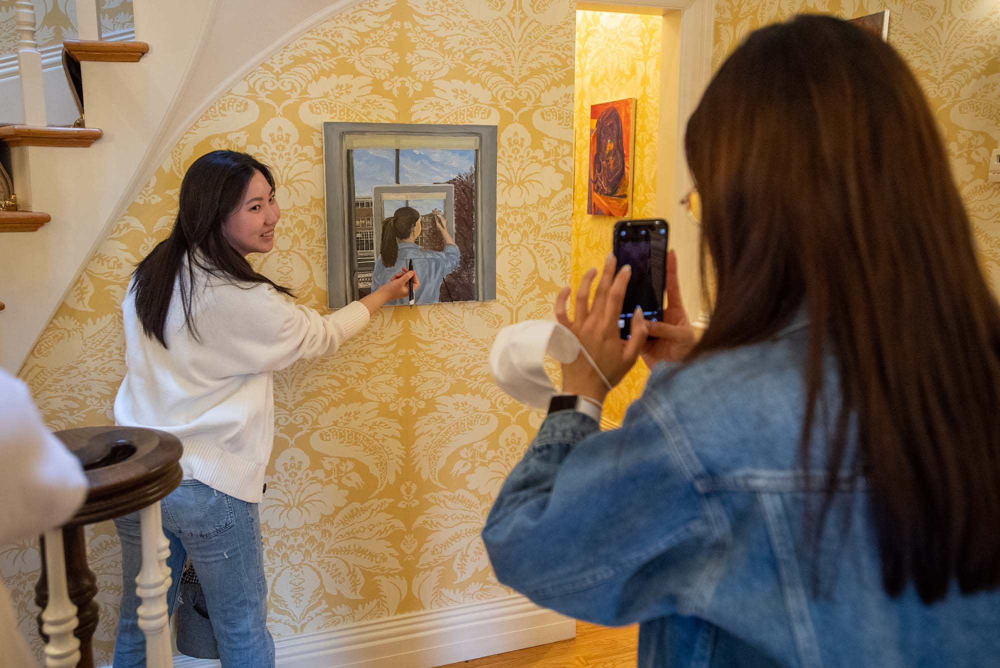Photo of Heeseung “Chelsea” Han (CFA’23), left, posing with her piece “Back to Me” while her friend Mina Chung (CFA’23) snaps a photo, right foreground, during the during the biannual art exhibit at Sloane House April 11. Han holds a pen and pretends to draw on her piece, which is an image of a person painting a boston skyline painting a boston skyline. The house has ornate, gold and white floral wallpaper. Han is a young, Asian woman with long dark hair and wears a white sweater and jeans.