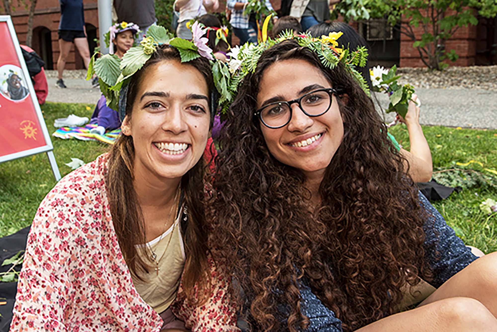 Photo of two young women with long brown hair smiling and wearing crowns made of green leaves. The woman on the right has curly hair and wears black framed glasses and the woman on the left has straight hair.