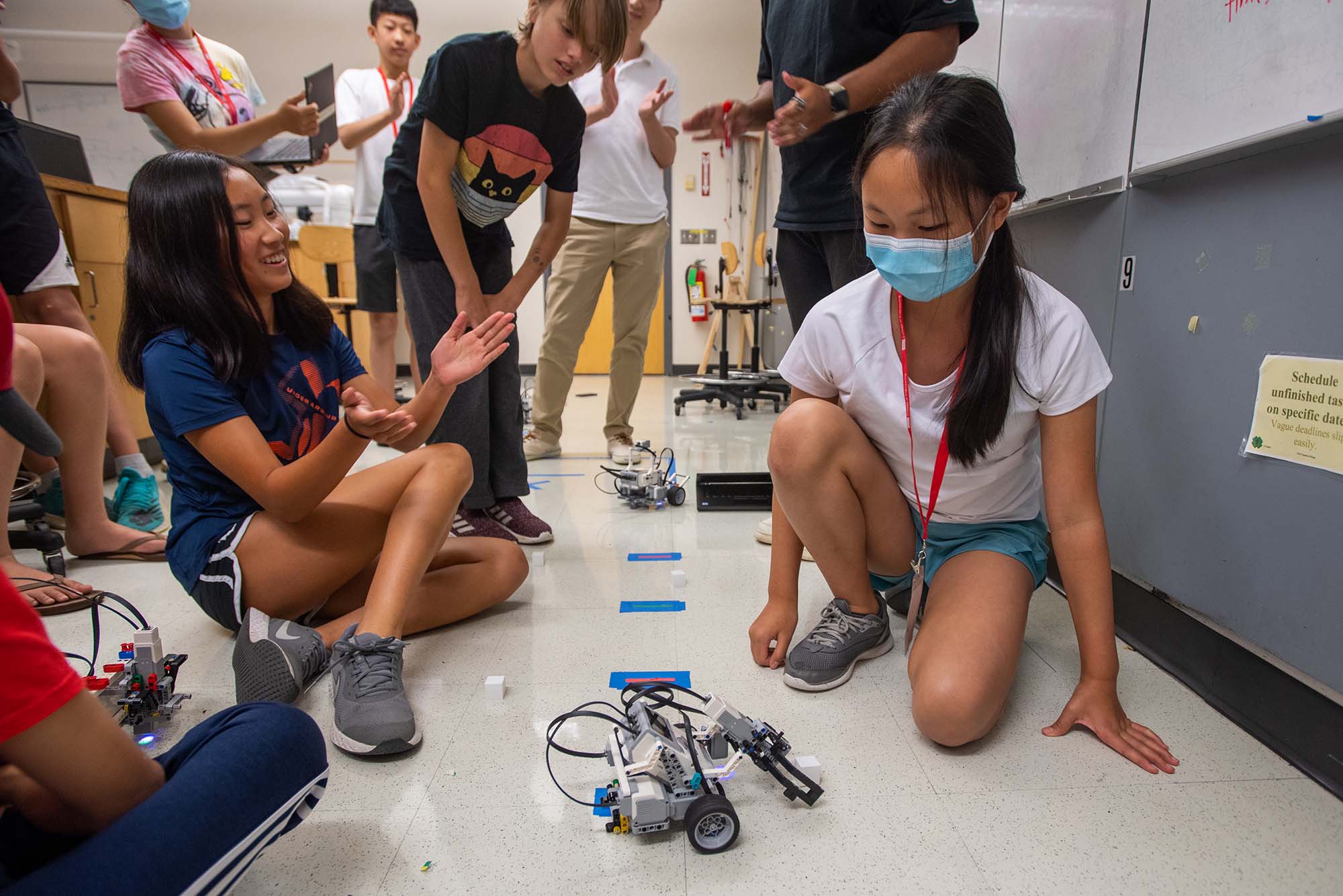 Photo of Raina Wu, 12, left, and Cindy Jin, 12, right, sigh with relief after their robot Marshmallow successfully performs a series of tasks after being programmed by the two along with their teammate Hailey Duan, 12, not pictured, in the U-Design Robo-Alley workshop at Photonics July 21. Jin kneels by the small gray and white robot that almost looks like it was built from legos in classroom; she is a young AAPI girl, and wears a white t-shirt, shorts, and a blue face mask. Wu, at left, smiles and claps her hands; she also sits on the ground. Wu is a young AAPI girl with long brown hair, a blue t-shirt, blue shorts, and sneakers on. Other students in the class are standing in the background, clapping.