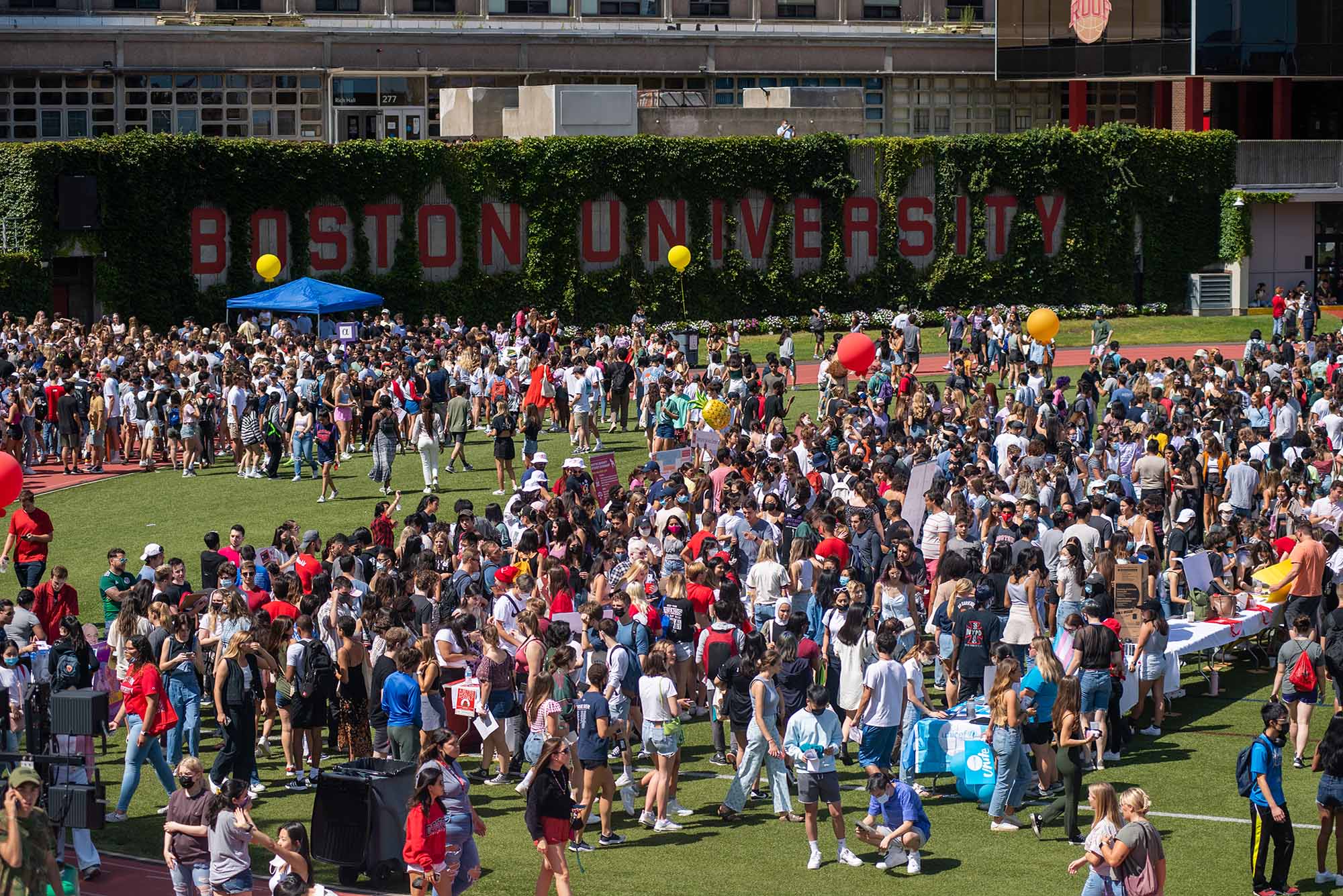 Photo of a crowd of BU students checking out booths, hanging around, eating, and just generally having fun on Nickerson Field, a soccer field with a track a red track surrounding it. The brick walls on one side of the field have large red letters that read "Boston University"