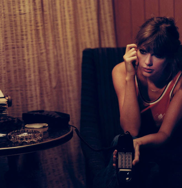 Image of Taylor Swift wearing an old school red tank top. She sits with black, corded phone in hand looking somber in a darker room.