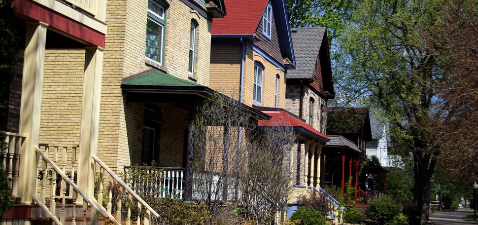 Photo: A sidewalk lined with cookie-cutter houses on the left side. They are tan with red, or blue roofs. Most have a porch with an awning on it. There are trees lining the other side of the sidewalk. The day is bright and sunny.