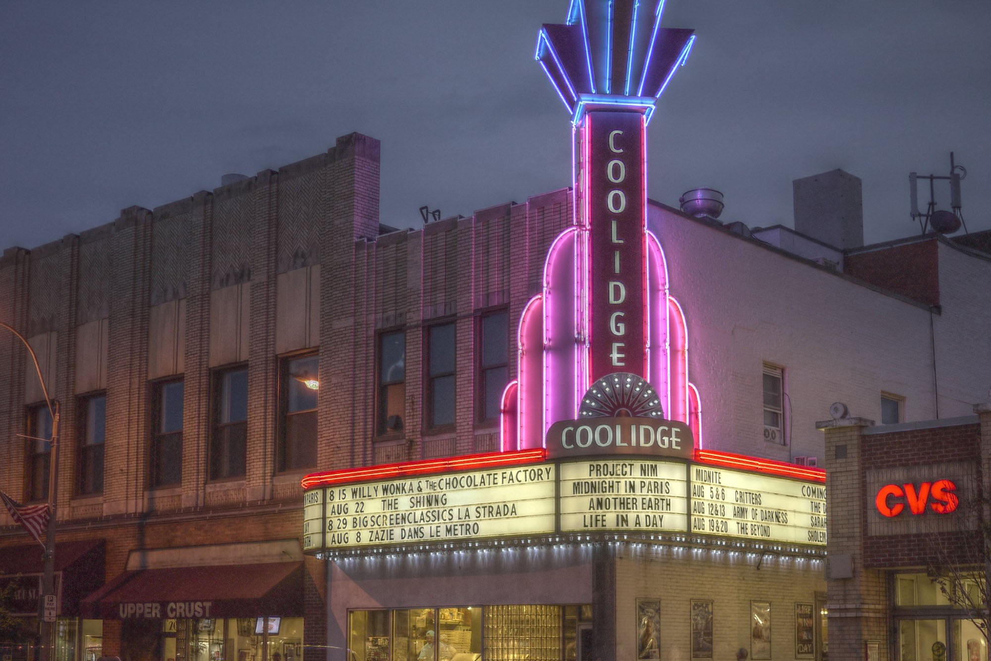 Photo: A brightly lit theater with blues and purples. In art deco style, the sign up top reads "COOLIDGE". Below the sign is a banner for the shows playing. The area is dark as its night tine.