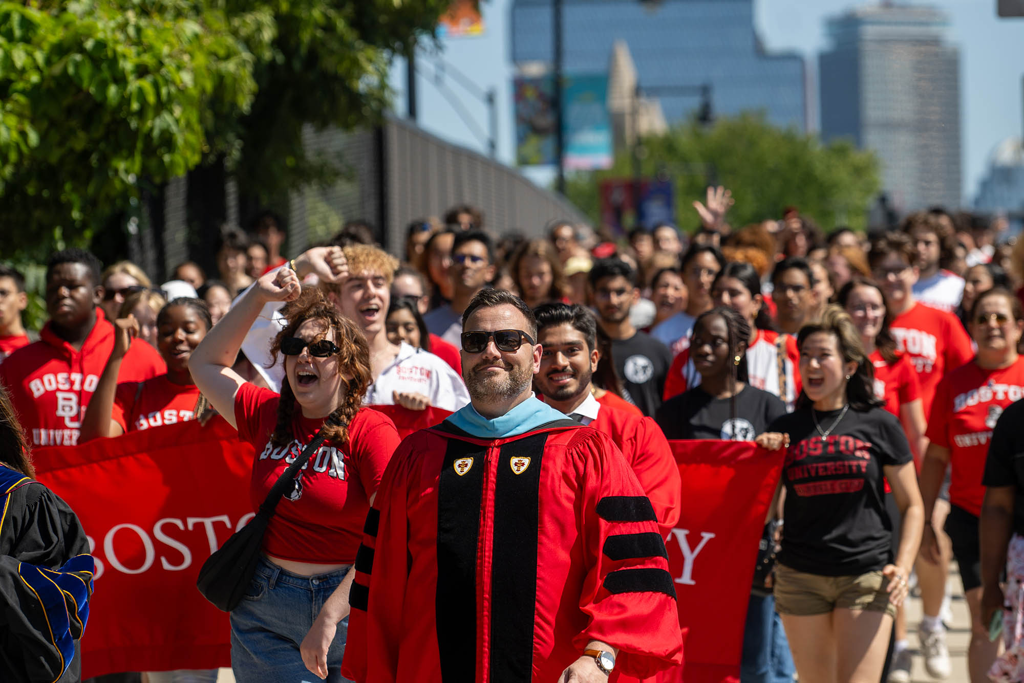 Photo: A large crowd of first year students and dean of students Jason Campbell-Foster parade up Commonwealth Ave. The dean of students wears sunglasses and a red ceremonial robe as he walks with a crowd of cheering, smiling students.