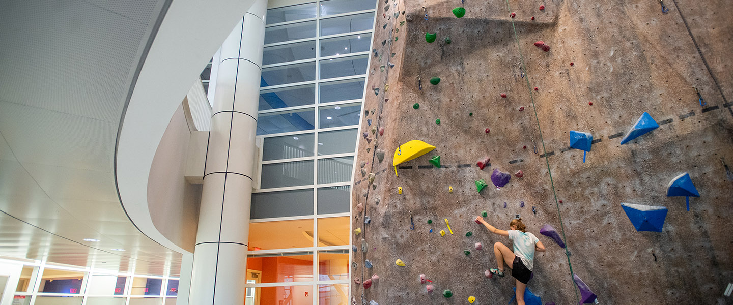 Photo of a young white woman with a light blue t shirt climbing on the rock climbing wall at fitrec. The wall is brownish gray and is a bit overhung; it's covered in colorful holds.