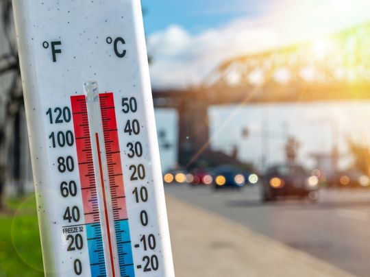 Thermometer reading 100 degrees fahrenheit with highway and cars in background.